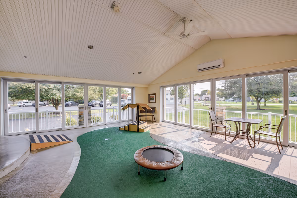 A bright indoor activity room with large windows and sliding glass doors providing a view of the outside greenery and parking area. The room features a small trampoline, a ramp, a wooden step platform, and a table with two chairs near the windows. The ceiling is white with recessed lighting and a ceiling fan, and there is an air conditioning unit mounted on the wall.