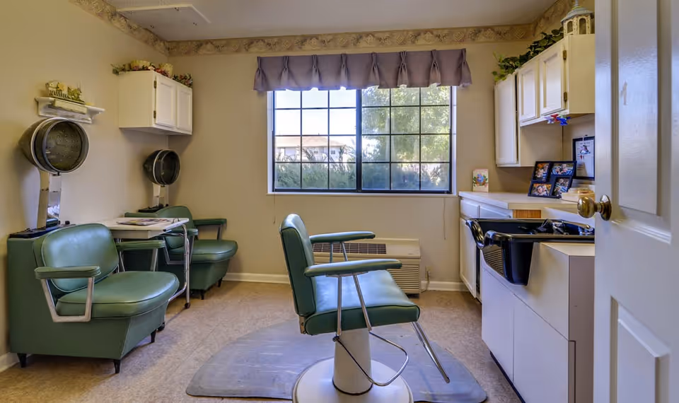 Interior of a small hair salon room with vintage green salon chairs, two hair dryers mounted on the wall, a large window with a purple valance, white cabinets, and a black salon sink. The room has beige walls with decorative border trim near the ceiling.