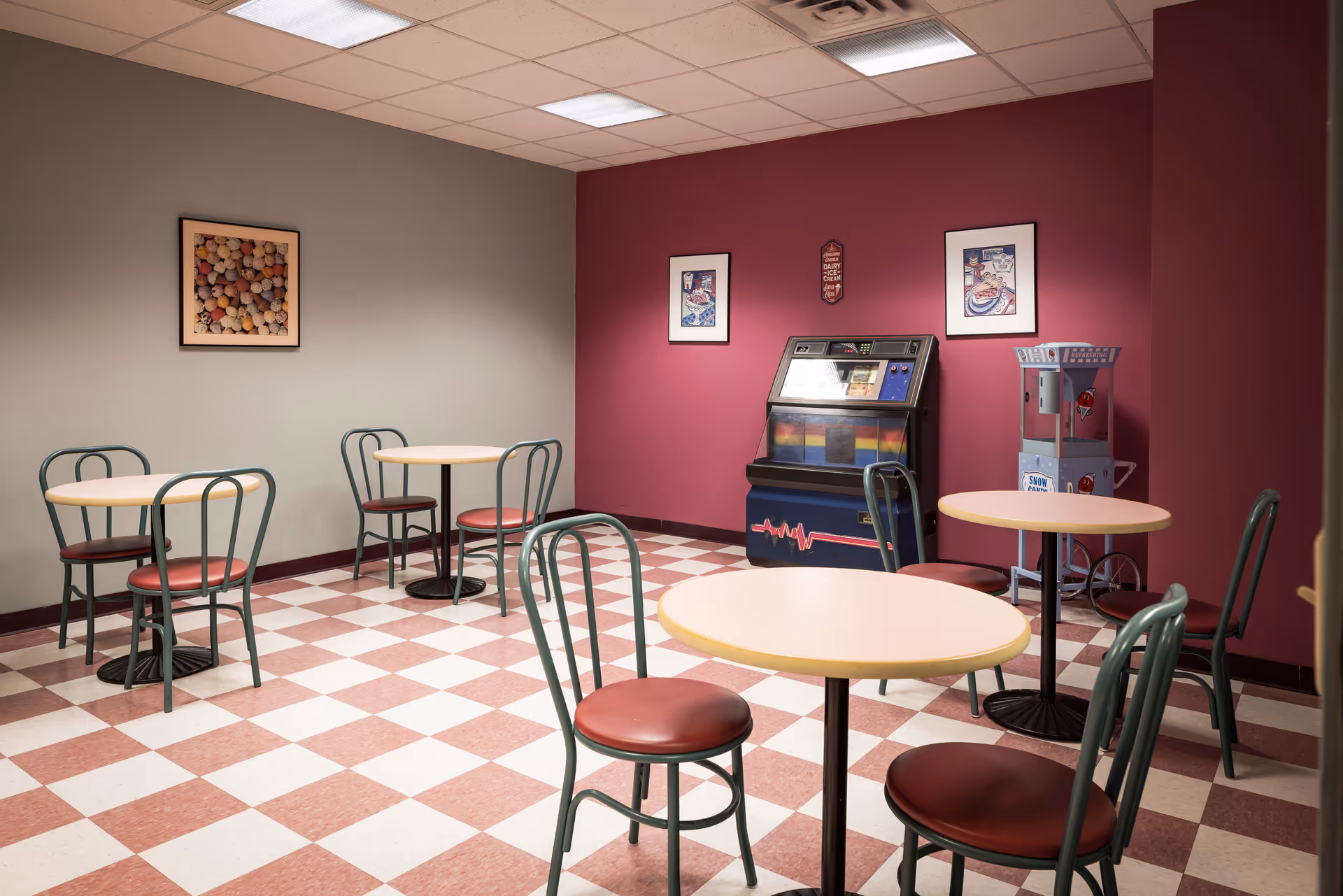 A small dining area with four round tables and green metal chairs with red cushions. The floor has a red and white checkered pattern. The walls are painted gray and dark red, with framed pictures and a vintage jukebox and popcorn machine against the red wall.