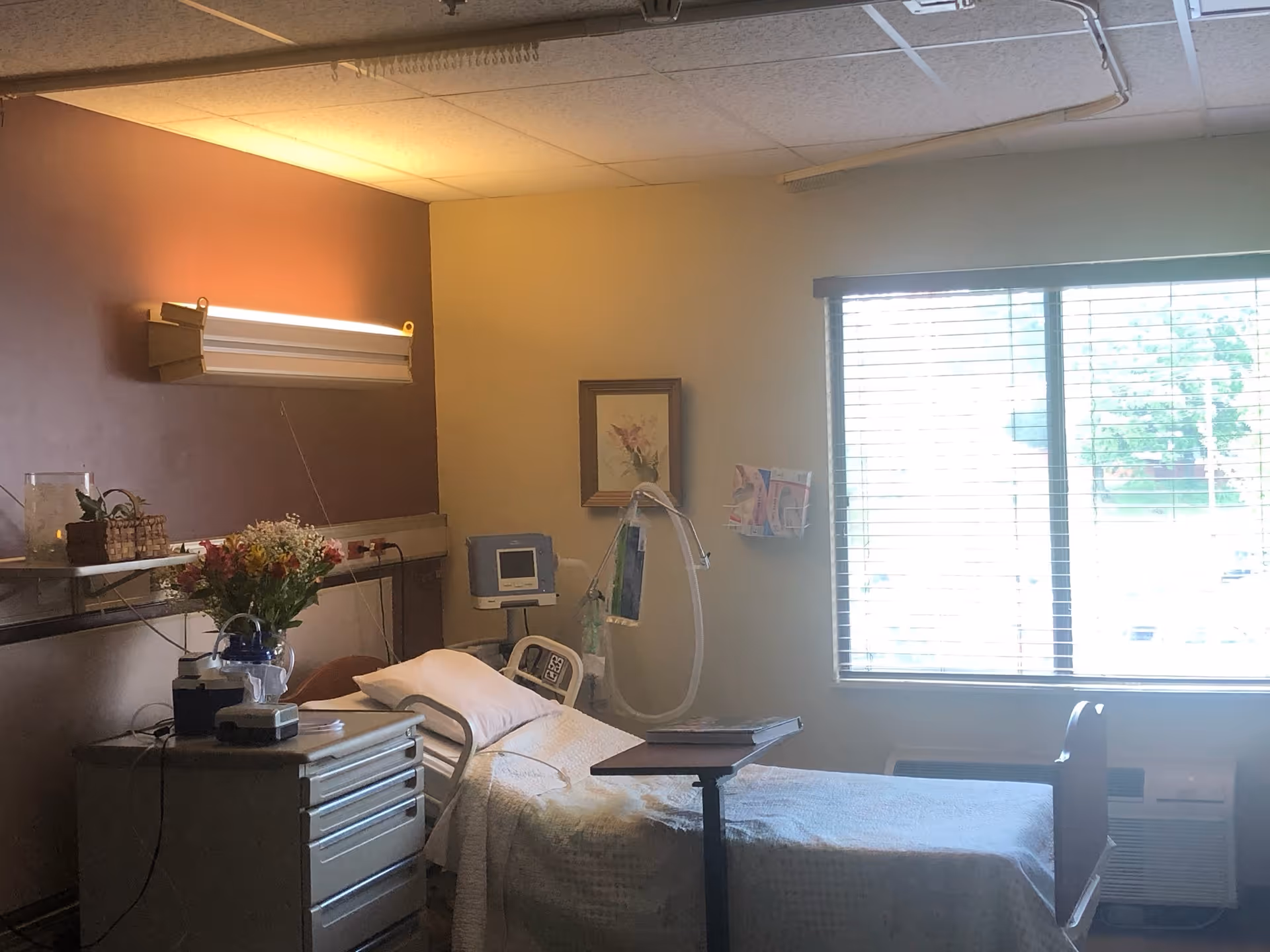 A hospital-style patient room with a single bed covered in white bedding, a bedside table with flowers and medical equipment, a wall-mounted light fixture, a framed picture on the wall, and a large window with blinds letting in natural light.