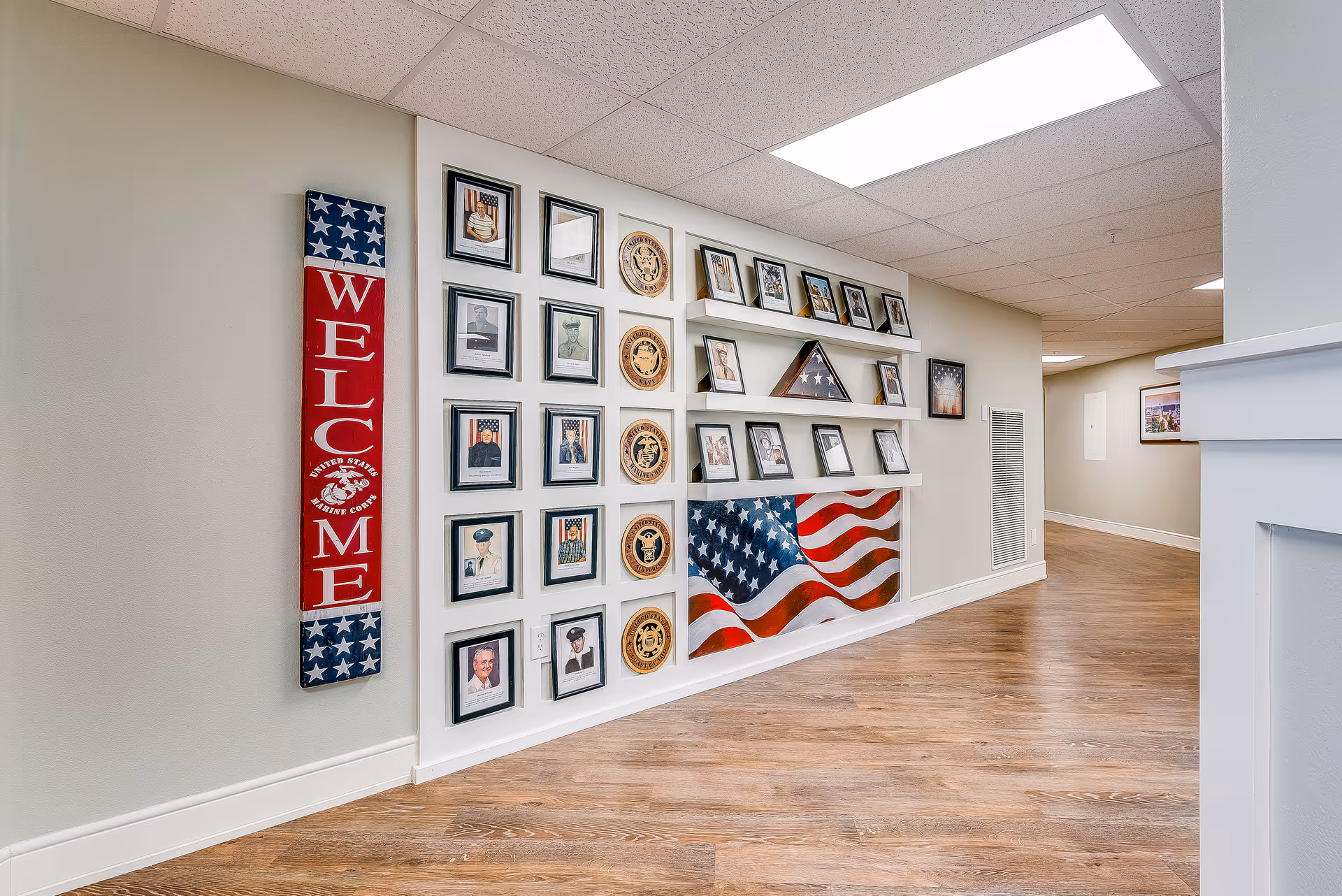 Interior hallway featuring a wall display of framed veteran photos, military emblems, an American flag mural, and a vertical 'WELCOME' sign.