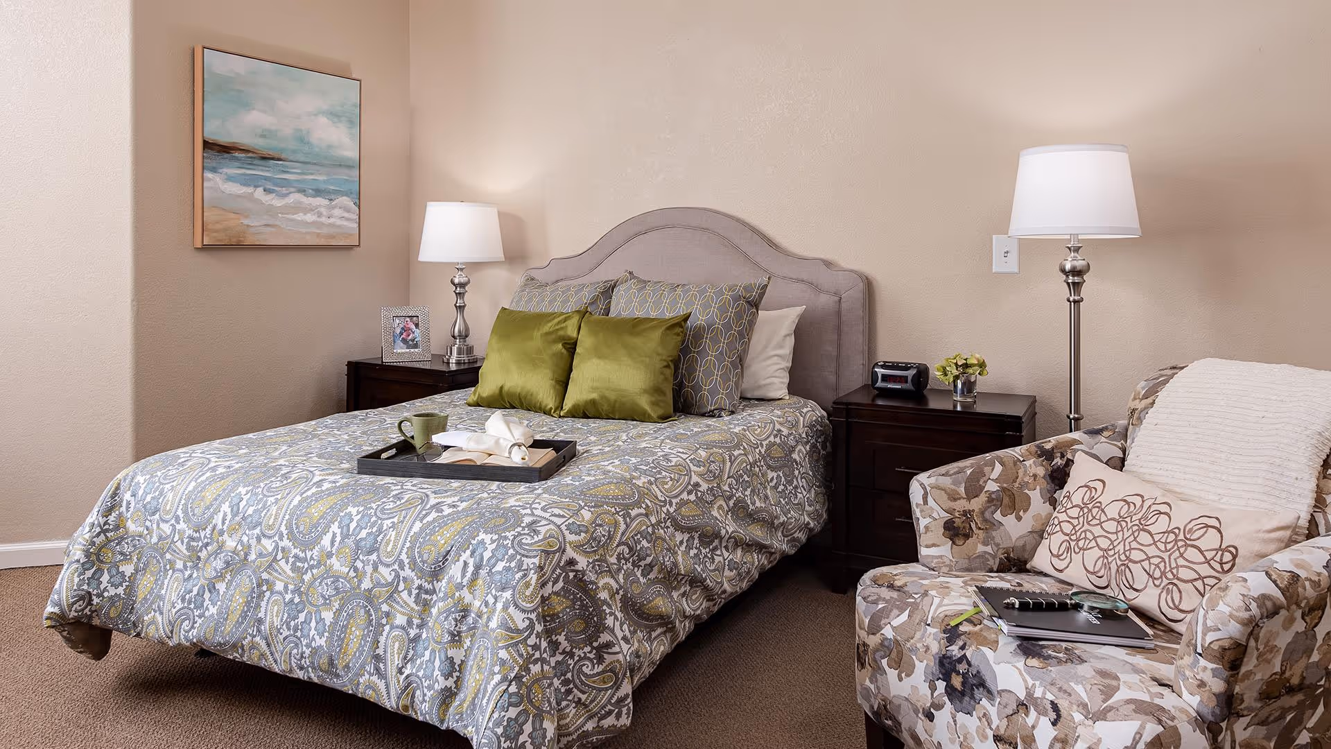 A cozy bedroom featuring a bed with a patterned bedspread and multiple pillows, two bedside tables each with a lamp, a floral armchair with a pillow and a notebook on it, and a framed beach painting on the wall.