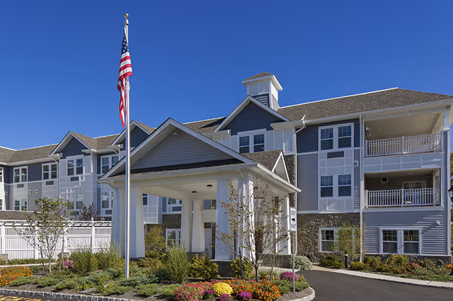 Exterior view of a senior living facility building with multiple windows and balconies under a clear blue sky. There is a covered entrance with white pillars and a flagpole with an American flag in front. The landscaping includes colorful flowers and shrubs along the driveway.