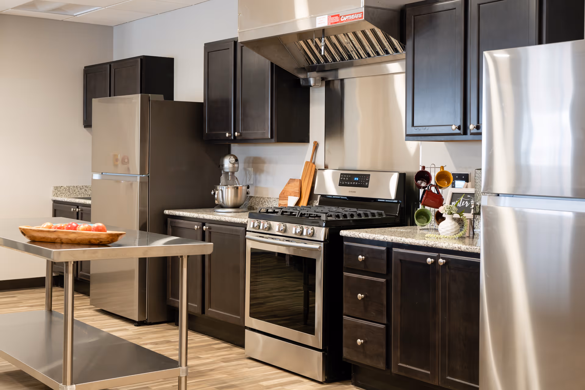 Modern communal kitchen with stainless steel appliances, dark wood cabinets, granite countertops, and a metal island with a bowl of apples.