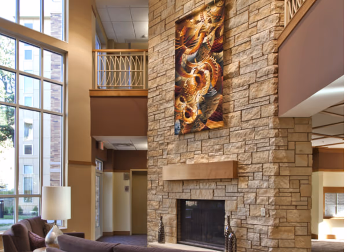 A spacious living room area with a tall stone fireplace as the focal point. Above the fireplace hangs a colorful abstract artwork. The room features large windows allowing natural light to fill the space, a brown sofa, and a white table lamp. There is a balcony railing visible on the upper level and warm beige and brown tones throughout the interior.