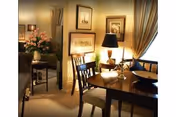 Cozy dining area with a wooden table and chairs, table lamp, framed artwork, and a floral arrangement.