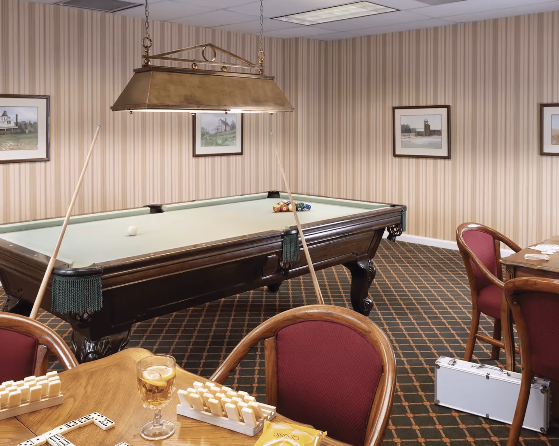 A recreational room with a pool table in the center, two pool cues resting against it, and a hanging light fixture above. The room has patterned carpet and striped wallpaper with framed pictures on the walls. In the foreground, there is a wooden table with dominoes, a glass of iced tea, and a pack of Classic potato chips. Several wooden chairs with red upholstery surround the table. A silver briefcase is on the floor near another table with playing cards.