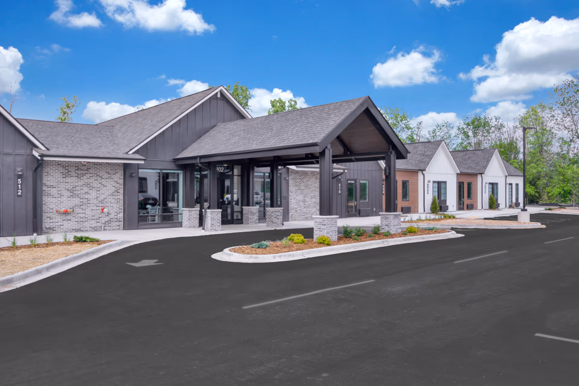 Front entrance of a senior living community building with a covered porte-cochere, landscaped islands and a paved driveway under a blue sky.