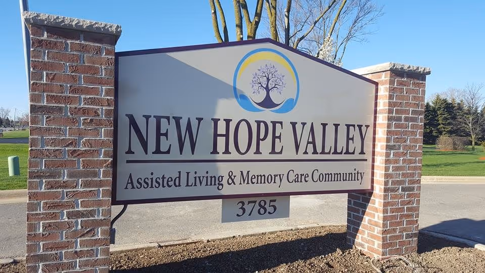 A large outdoor sign for New Hope Valley Assisted Living & Memory Care Community, mounted between two brick pillars with a clear blue sky and green grass in the background.
