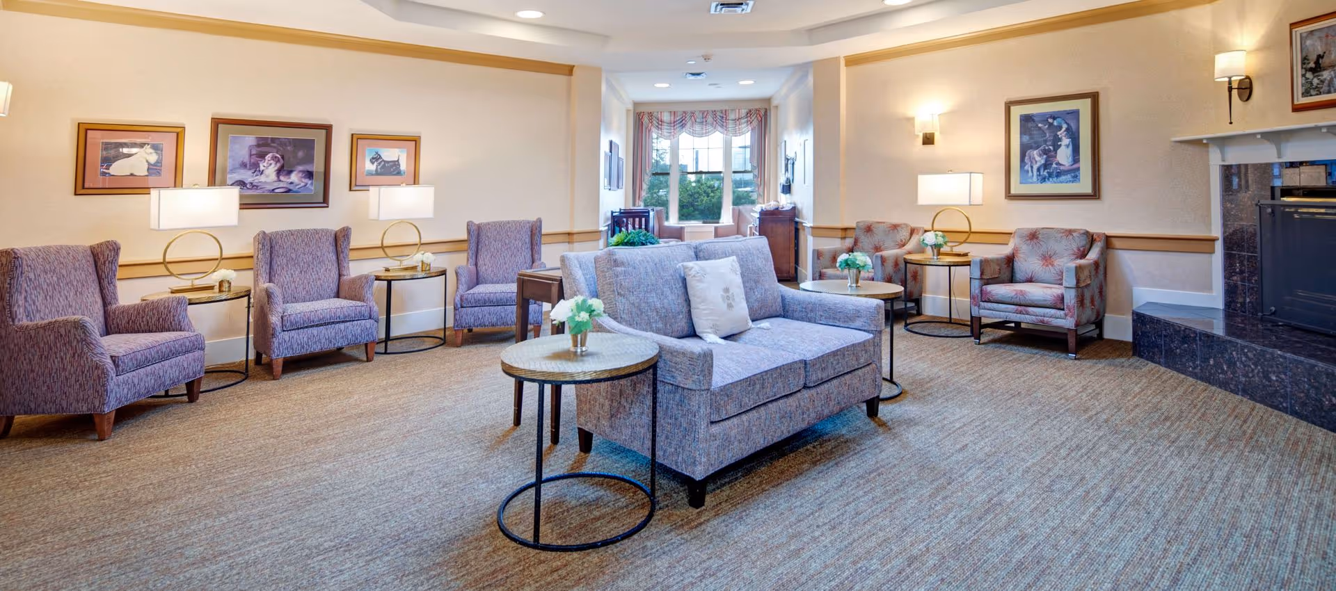 A cozy living room area in a senior living facility with multiple upholstered chairs and a loveseat arranged around small round tables with lamps and flower vases. The room has beige walls adorned with framed pictures, a carpeted floor, and a fireplace with a dark marble surround. A window with patterned curtains lets in natural light at the far end of the room.