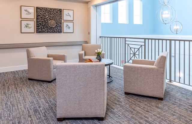 A bright and modern sitting area with four beige upholstered armchairs arranged around a round coffee table with books and a small vase of flowers. The space has large windows with metal railings allowing natural light to fill the room. On the wall, there are five framed botanical artworks arranged in a grid. The floor is carpeted with a textured pattern.