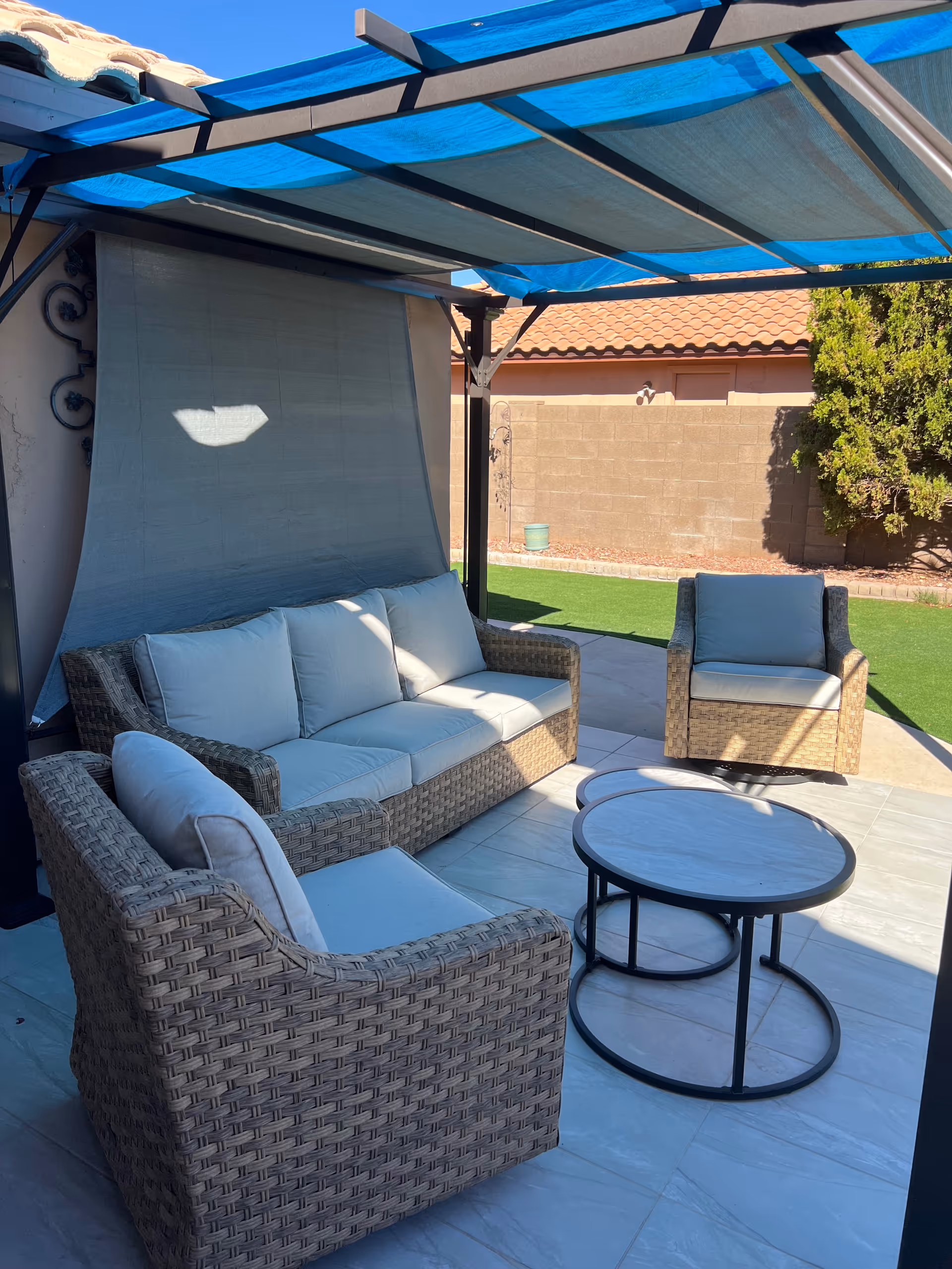 Outdoor patio area with a wicker sofa and two matching armchairs with light cushions, a round coffee table, and a blue fabric canopy providing shade. The patio is tiled and adjacent to a lawn with a brick wall and some greenery in the background.