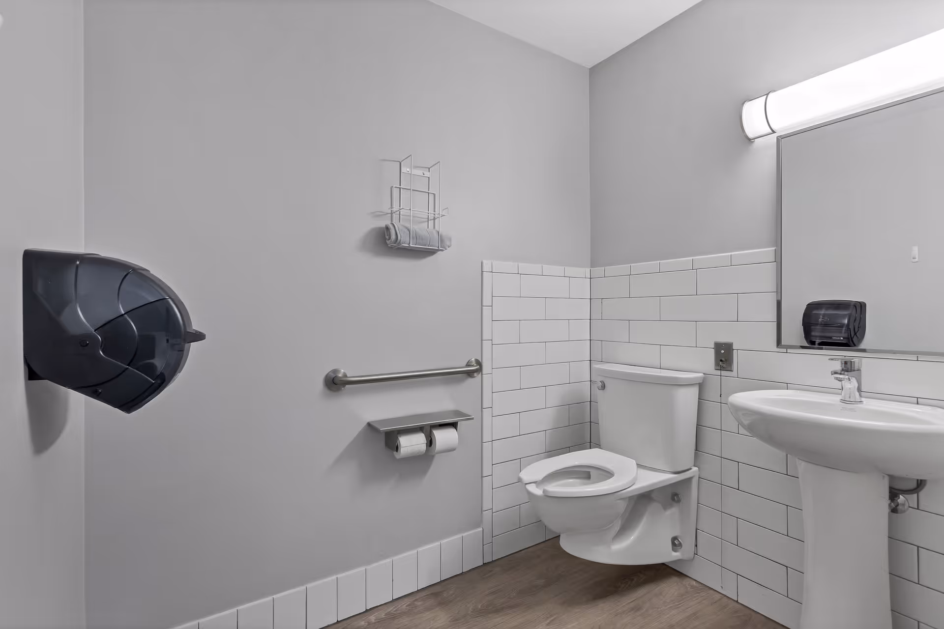 A clean, accessible bathroom with a white toilet, white pedestal sink, large mirror, and a wall-mounted handrail. The walls are painted light gray with white subway tile halfway up. There is a black paper towel dispenser on the left wall and a towel rack with a rolled towel above the handrail.