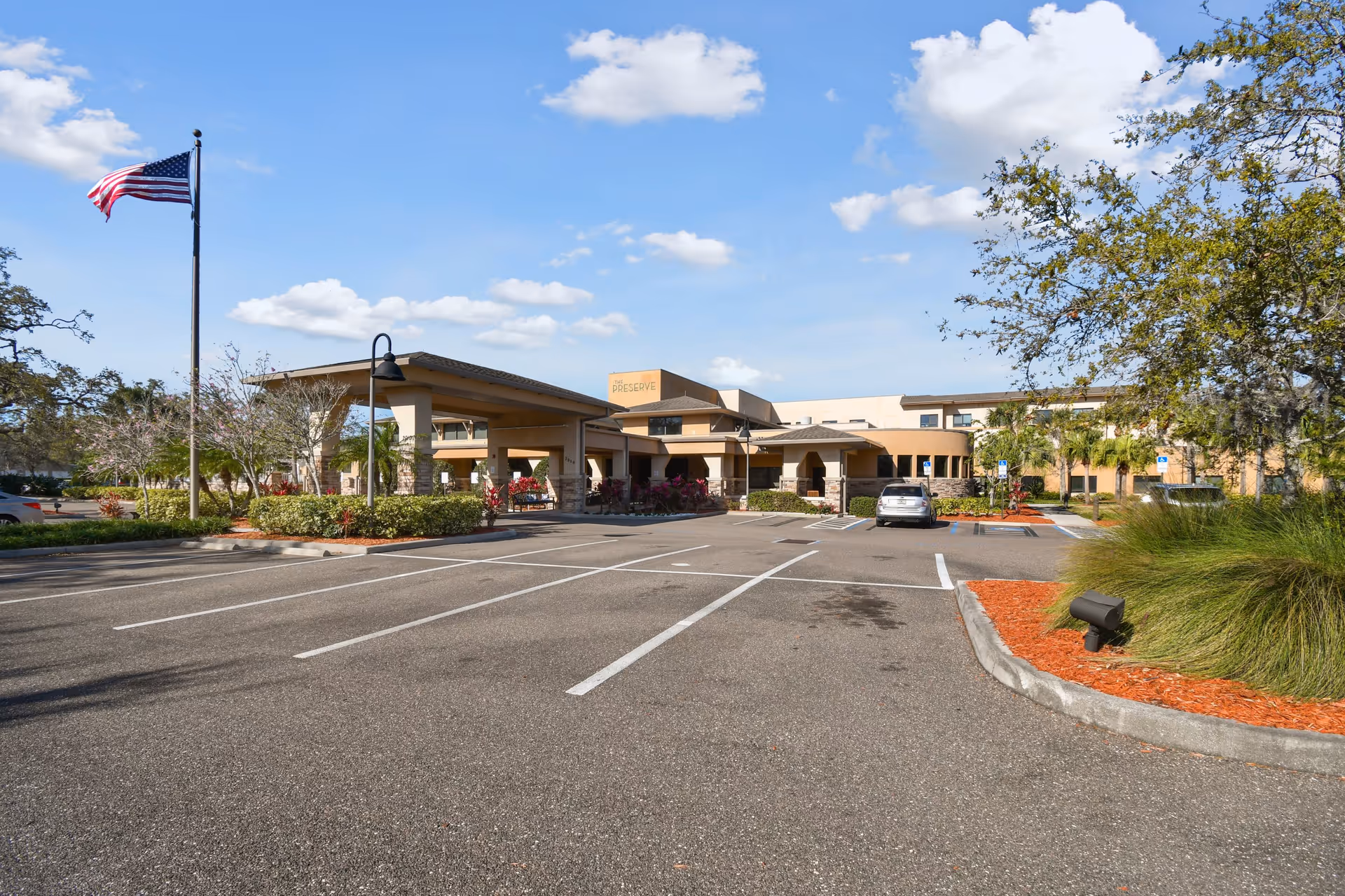Exterior view of The Preserve at Dunedin senior living facility with a parking lot in front, an American flag on a flagpole, landscaped greenery, and a partly cloudy blue sky.