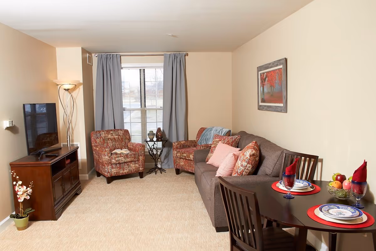 A cozy living room with a gray sofa adorned with patterned and solid pink pillows, two patterned armchairs, a small round glass table with decorative items between the chairs, a wooden TV stand with a flat-screen TV, a tall floor lamp, and a dining table set for two with plates, glasses, red napkins, and a bowl of fruit. Light beige walls and carpet, with a window covered by light blue curtains.