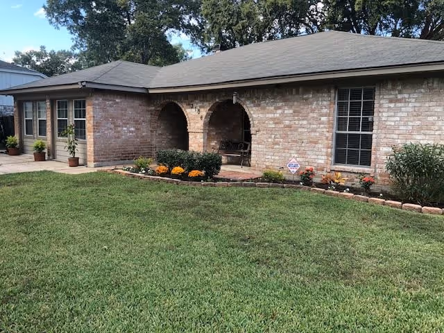 Single-story brick building with a covered front porch featuring two arches, a well-maintained green lawn, and a flower bed with various plants and flowers along the front. Trees are visible in the background under a partly cloudy sky.