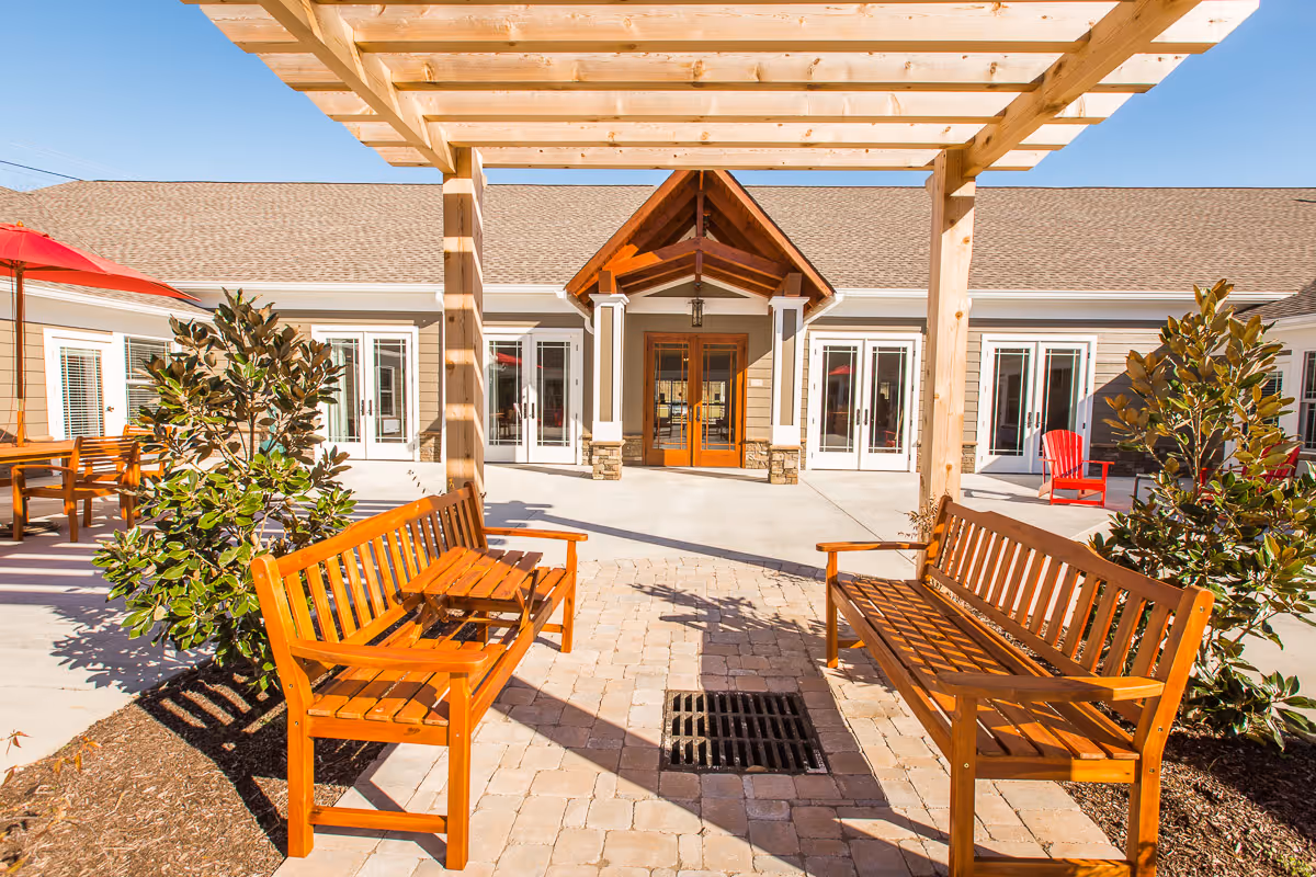 Outdoor seating area at Signature Lifestyles of Jefferson City featuring two wooden benches facing each other under a wooden pergola, with a paved stone floor and plants on either side. In the background, there is a building with multiple glass doors and a peaked wooden entrance.
