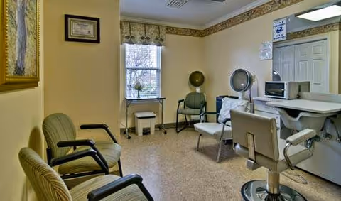 A small hair salon room with two salon chairs, a hair dryer hood, a microwave on a counter, and a window with a floral valance. There are three green chairs along one wall and framed pictures hanging on the walls.