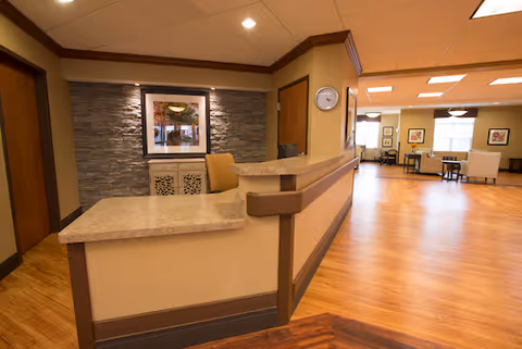 Reception desk area with a stone accent wall behind it, a framed picture, and a clock on the wall. The space has wood flooring and leads into a well-lit common area with tables and chairs.