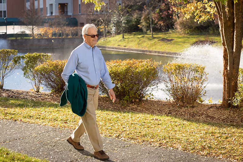 An elderly man wearing sunglasses, a light blue shirt, beige pants, and brown shoes walks along a paved path beside a pond with a water fountain. He is holding a green jacket in his left hand. The background includes bushes, trees, and a building partially visible behind the pond.