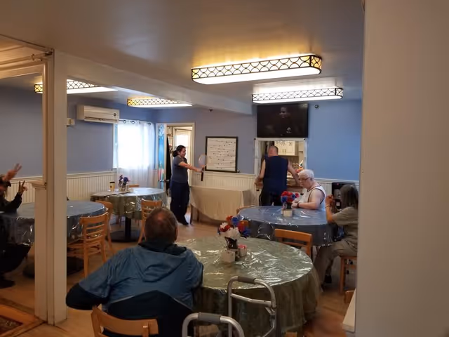Several residents and staff sit and stand around round tables in a communal dining/activity room with plastic-covered tablecloths.