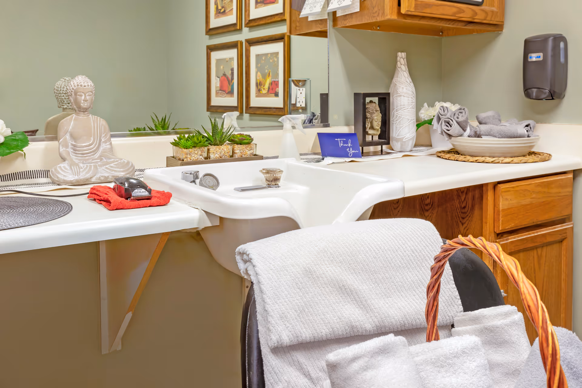 Well-decorated bathroom vanity with a white sink, folded towels and a basket, decorative Buddha statue and plants in front of a mirror.