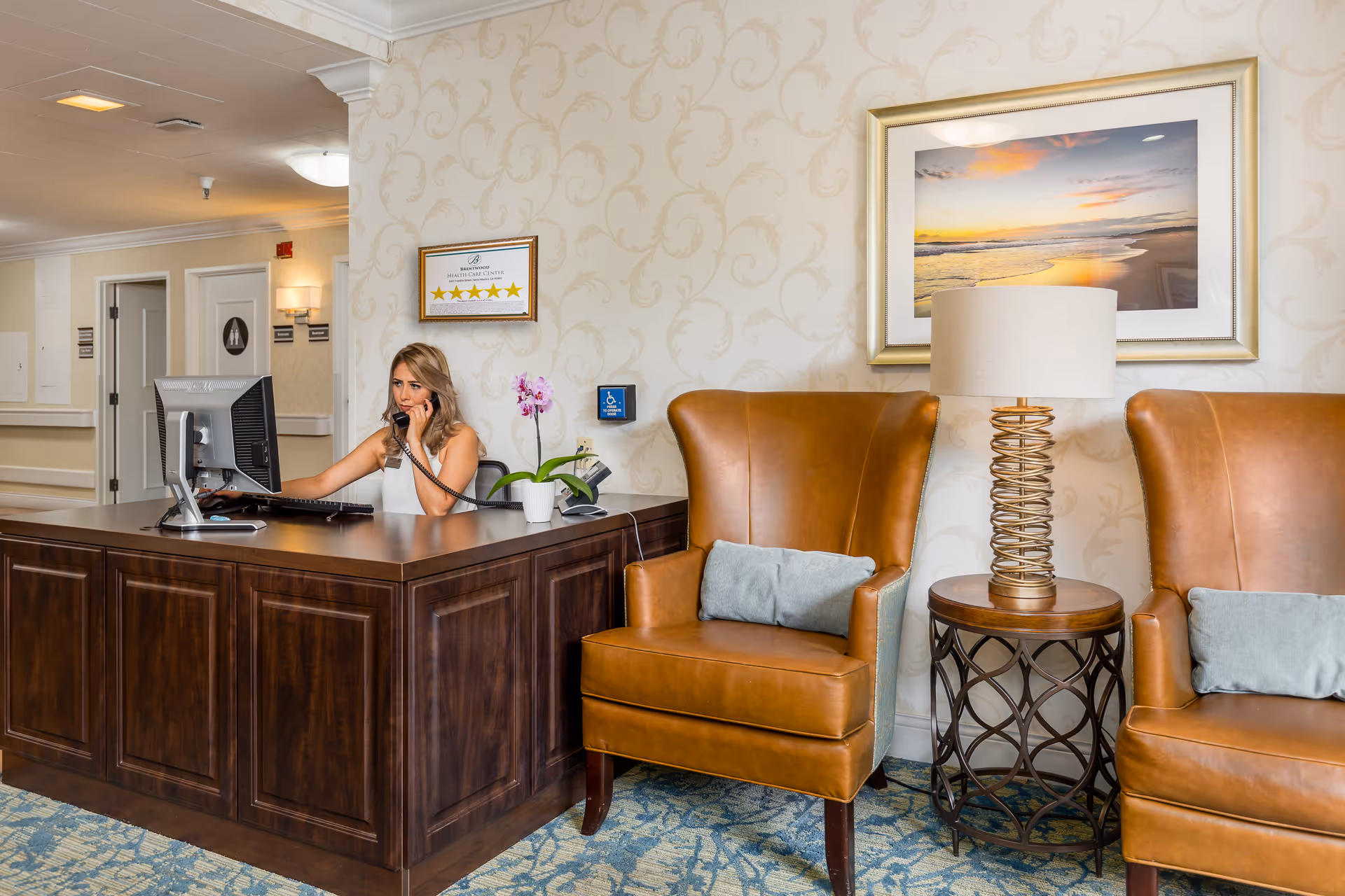 Reception area of Brentwood Health Care Center with a woman sitting behind a dark wooden desk, talking on the phone and using a computer. The area features two brown leather armchairs with gray cushions, a round side table with a decorative lamp, and a framed picture of a beach sunset on the wall. The wallpaper has a subtle floral pattern, and a framed certificate with five stars is displayed behind the desk.