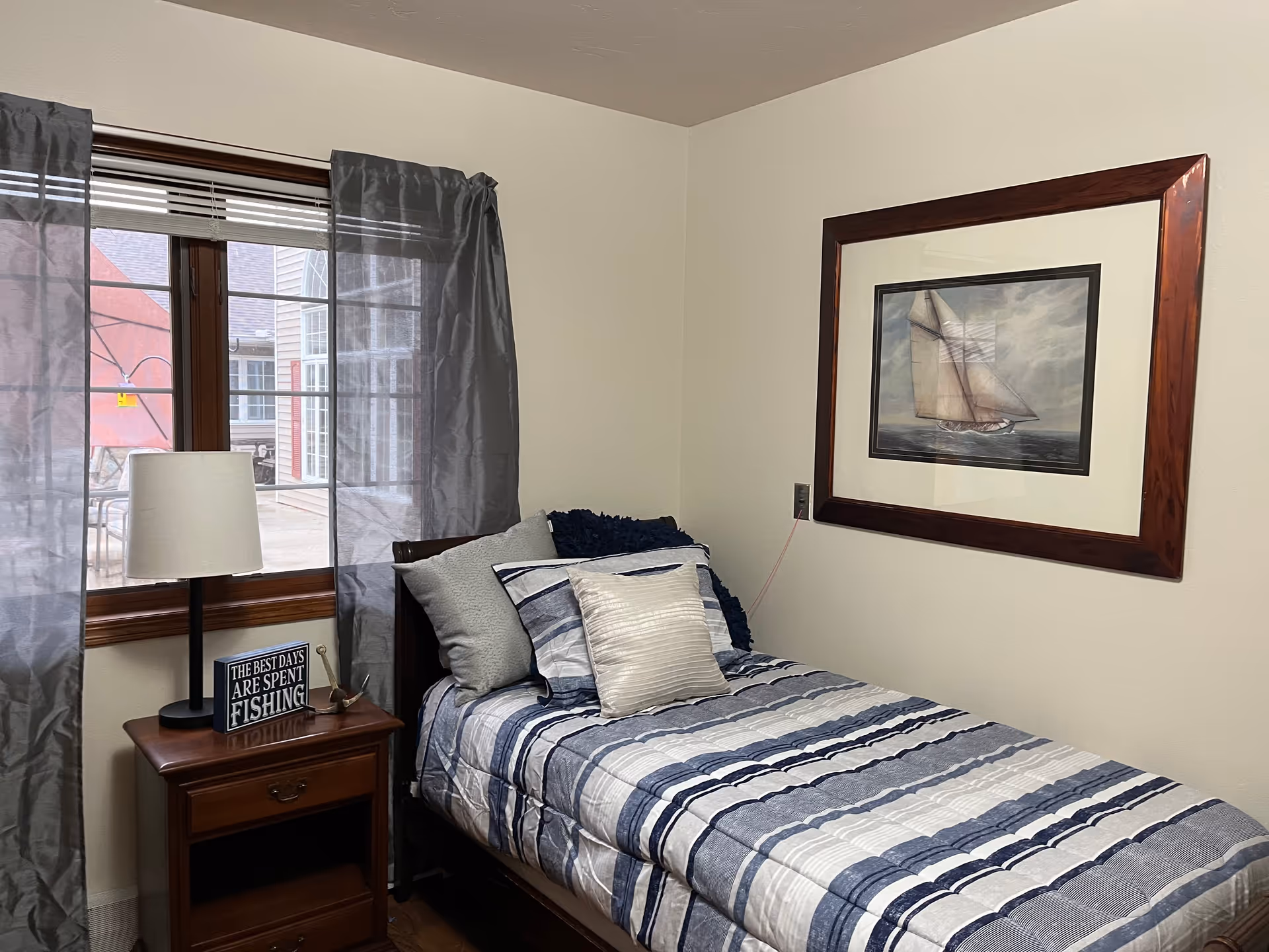 A cozy bedroom with a single bed covered in a blue, white, and gray striped comforter and several pillows. Next to the bed is a wooden nightstand with a lamp and a decorative sign that reads 'THE BEST DAYS ARE SPENT FISHING'. A window with gray curtains is behind the nightstand, and a framed picture of a sailboat hangs on the wall above the bed.