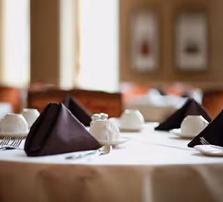 A close-up view of a dining table set with white tablecloth, folded dark brown napkins, white cups, and silverware in a softly lit dining room with blurred background.
