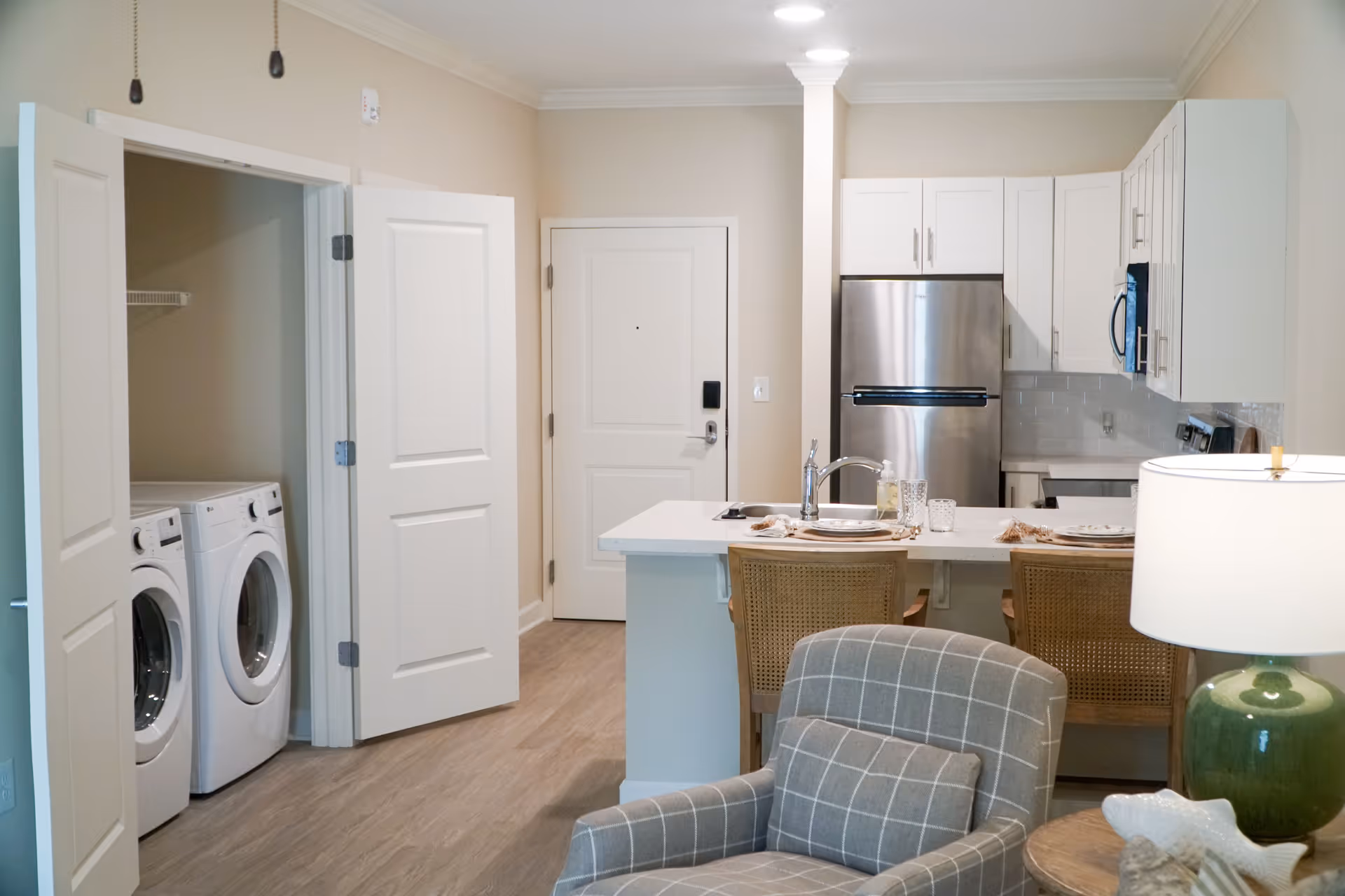 Open-plan apartment interior showing a kitchen with an island and bar stools, a washer/dryer closet, and a plaid armchair in the foreground.