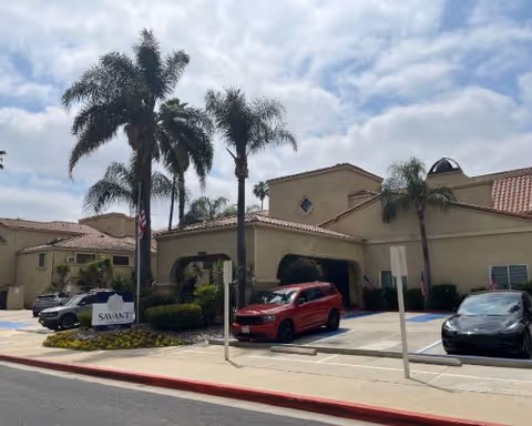 Beige senior living facility front with arches, palm trees, and parked cars.