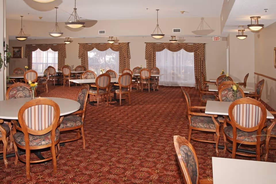 A spacious dining room with multiple round and square tables surrounded by wooden chairs with patterned upholstery. The room has large windows with decorative curtains, a red patterned carpet, and ceiling lights evenly spaced throughout.