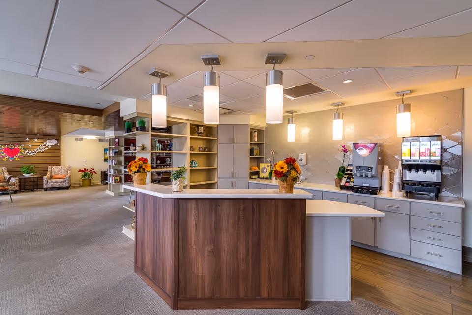 Interior view of a senior living facility's common area featuring a reception or service counter with hanging pendant lights above. The counter has flower arrangements and small plants. Behind the counter, there are beverage dispensers and cabinets. In the background, there are shelves with decorative items and a seating area with armchairs and wall decorations.