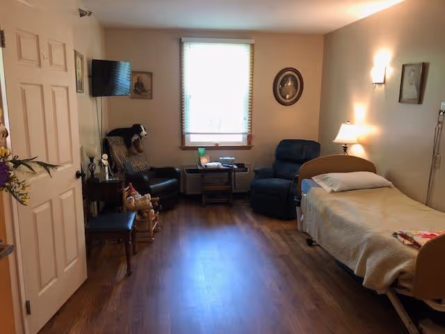 A cozy room with a single bed on the right side covered with a beige blanket and a pillow. Next to the bed is a small lamp on a nightstand. Two comfortable armchairs are positioned near a window with blinds, and a small table with items on it is between the chairs. The floor is wooden, and the walls have framed pictures and a wall-mounted TV. A door is partially open on the left side, and there are some decorative items including a rocking horse and flowers.