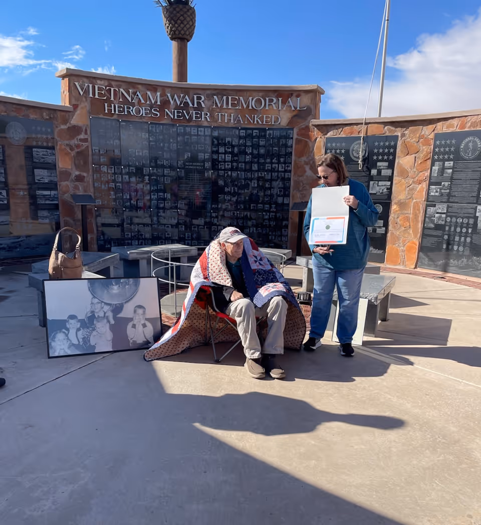 An elderly man sitting in a folding chair wrapped in a quilt at the Vietnam War Memorial outdoors. A woman stands next to him holding a certificate or document. Behind them is a stone wall with plaques and the inscription 'Vietnam War Memorial Heroes Never Thanked'. A large black and white photo of children is propped against a bench nearby.