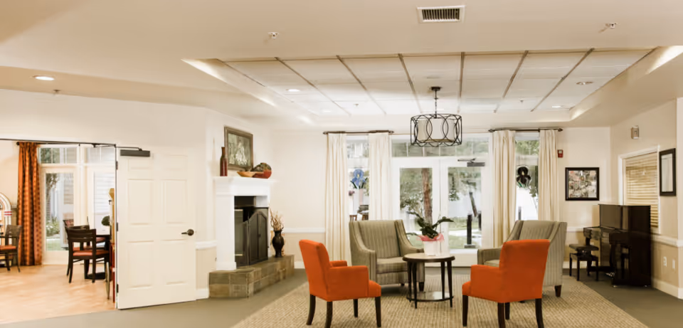 A cozy living room area in a senior living facility with two orange armchairs and two gray armchairs arranged around a small round table with a potted plant. There is a fireplace with decorative items on the mantel, a piano against the wall, and large windows with white curtains letting in natural light. A dining area with tables and chairs is visible through an open doorway on the left.