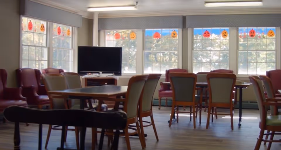 A bright common area in a senior living facility with multiple tables and chairs arranged for group seating. There are large windows decorated with hanging Halloween pumpkin decorations, allowing natural light to fill the room. A television is placed against the wall near the windows, and several comfortable armchairs are positioned along the walls.