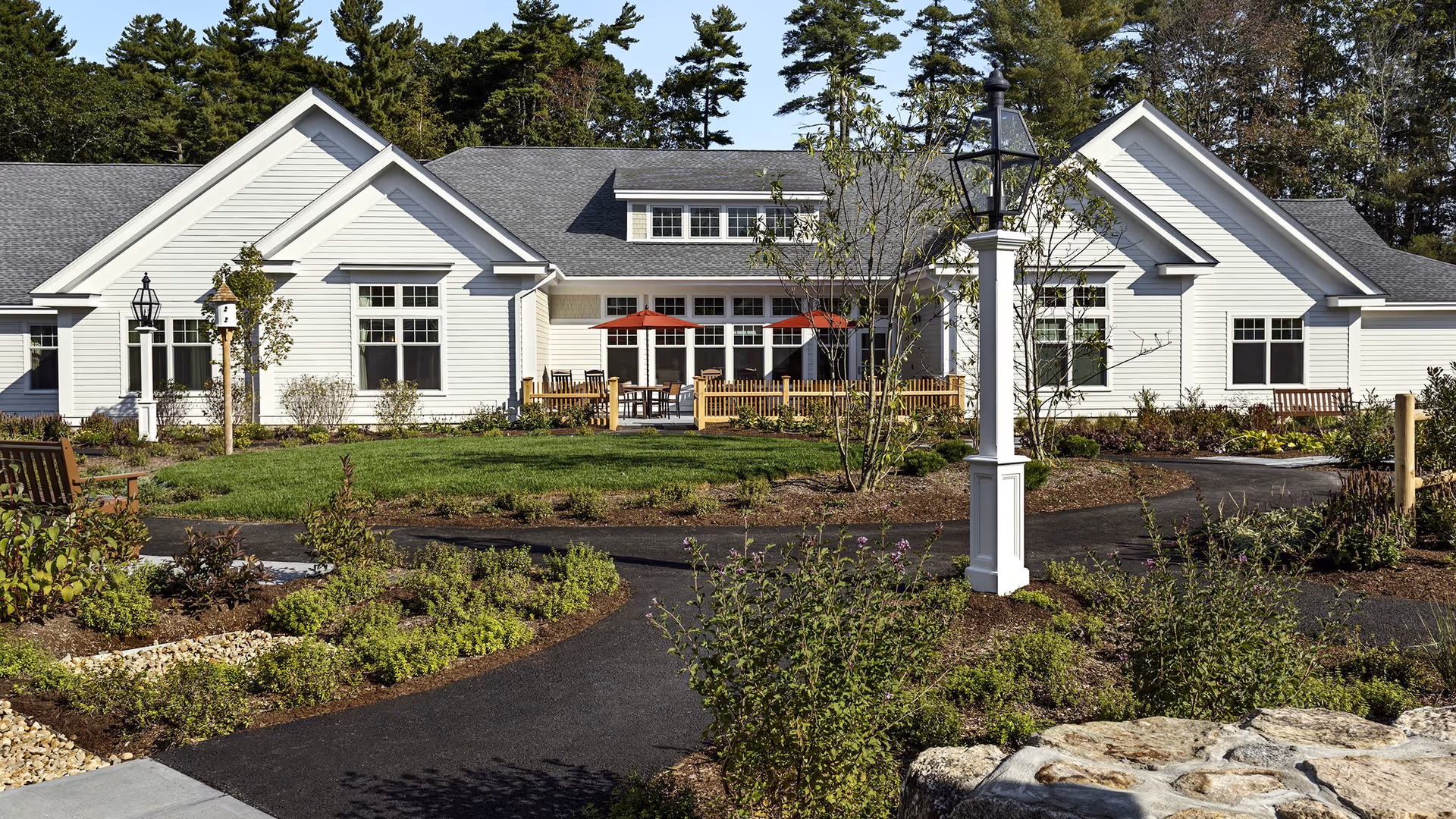 Exterior view of a white single-story building with a gray roof, surrounded by landscaped gardens and paved walkways. There are benches, a birdhouse, and outdoor seating with red umbrellas on a fenced patio. Tall trees are visible in the background under a clear sky.