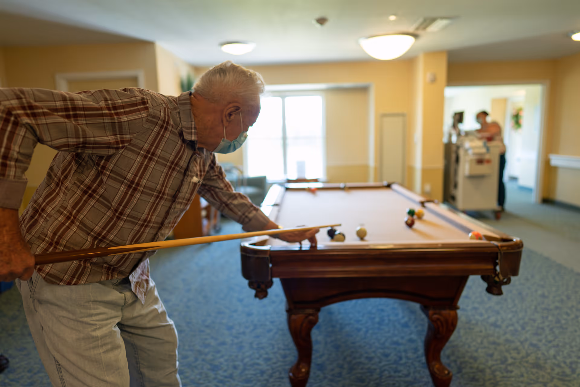 An elderly man wearing a face mask and a plaid shirt is playing pool indoors, aiming with a cue stick at a billiard table. The room has carpeted floors and beige walls, with two people in the background near a doorway.