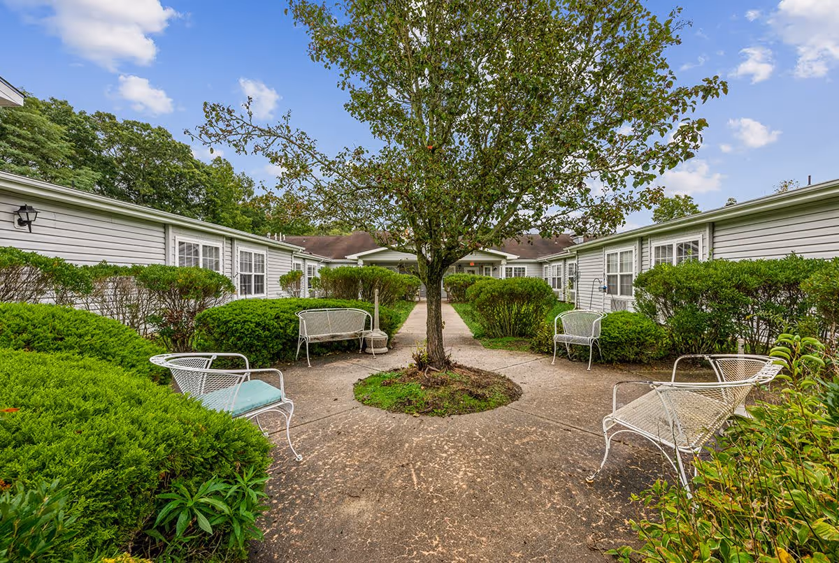 Outdoor courtyard area at The Addison of Somers Place featuring a central tree surrounded by a circular concrete path with white metal benches and green bushes. The courtyard is enclosed by single-story buildings with white siding and multiple windows under a partly cloudy blue sky.