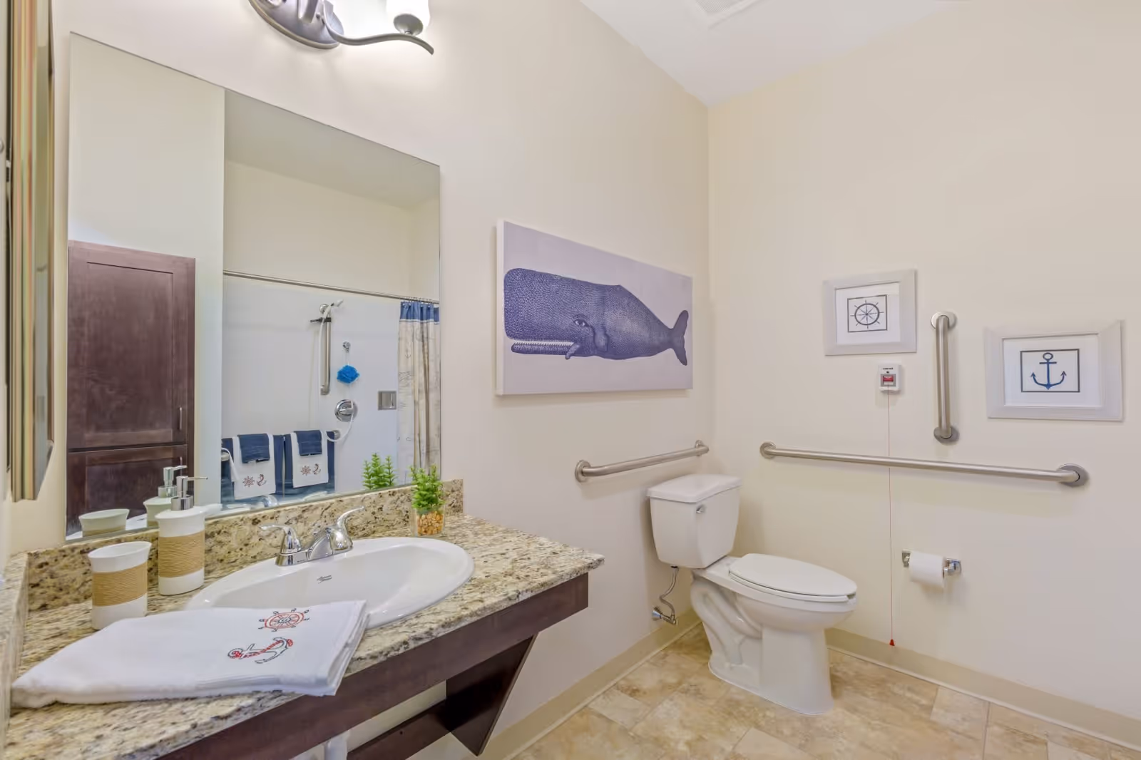 A clean and accessible bathroom with a granite countertop sink, a white toilet with grab bars on the walls, nautical-themed wall art including a whale, an anchor, and a ship's wheel, and a towel with an embroidered anchor on the counter.