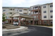 Exterior view of a multi-story senior living facility named Lucy Corr, featuring a covered entrance with a driveway and parking area in front. The building has a combination of brick and light-colored siding with multiple windows and balconies.