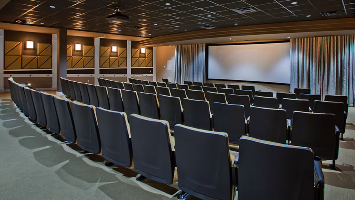 A theater-style room with multiple rows of black cushioned chairs facing a large blank projection screen. The walls have decorative panels and wall-mounted lights, and the ceiling has a grid pattern with recessed lighting.