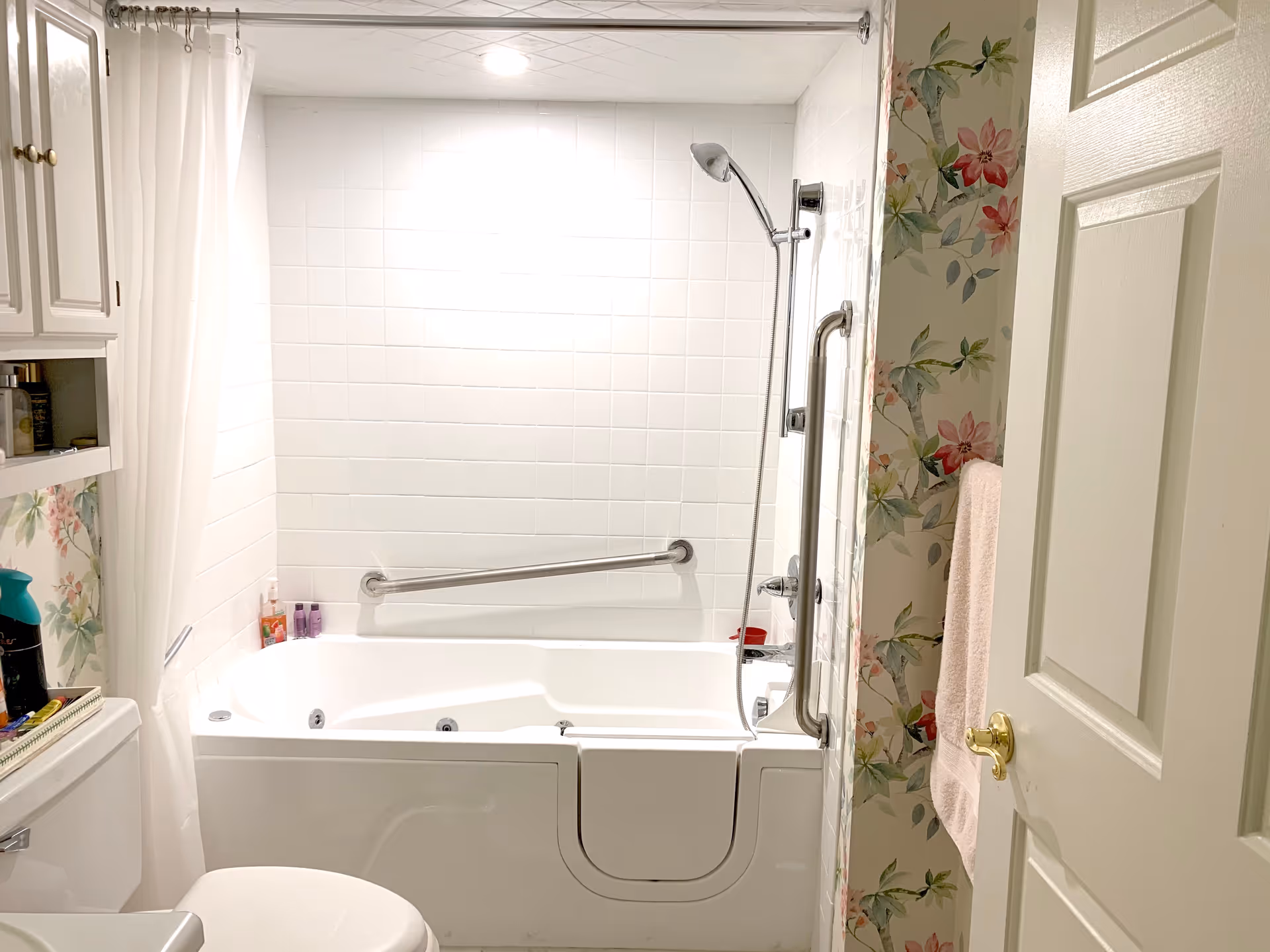 A bright bathroom featuring a walk-in bathtub with a safety grab bar and a handheld showerhead. The walls are tiled in white, and there is floral wallpaper on the adjacent wall. A white shower curtain hangs on a rod above the tub. A white toilet is partially visible in the foreground, and a towel hangs on a rack next to the door.