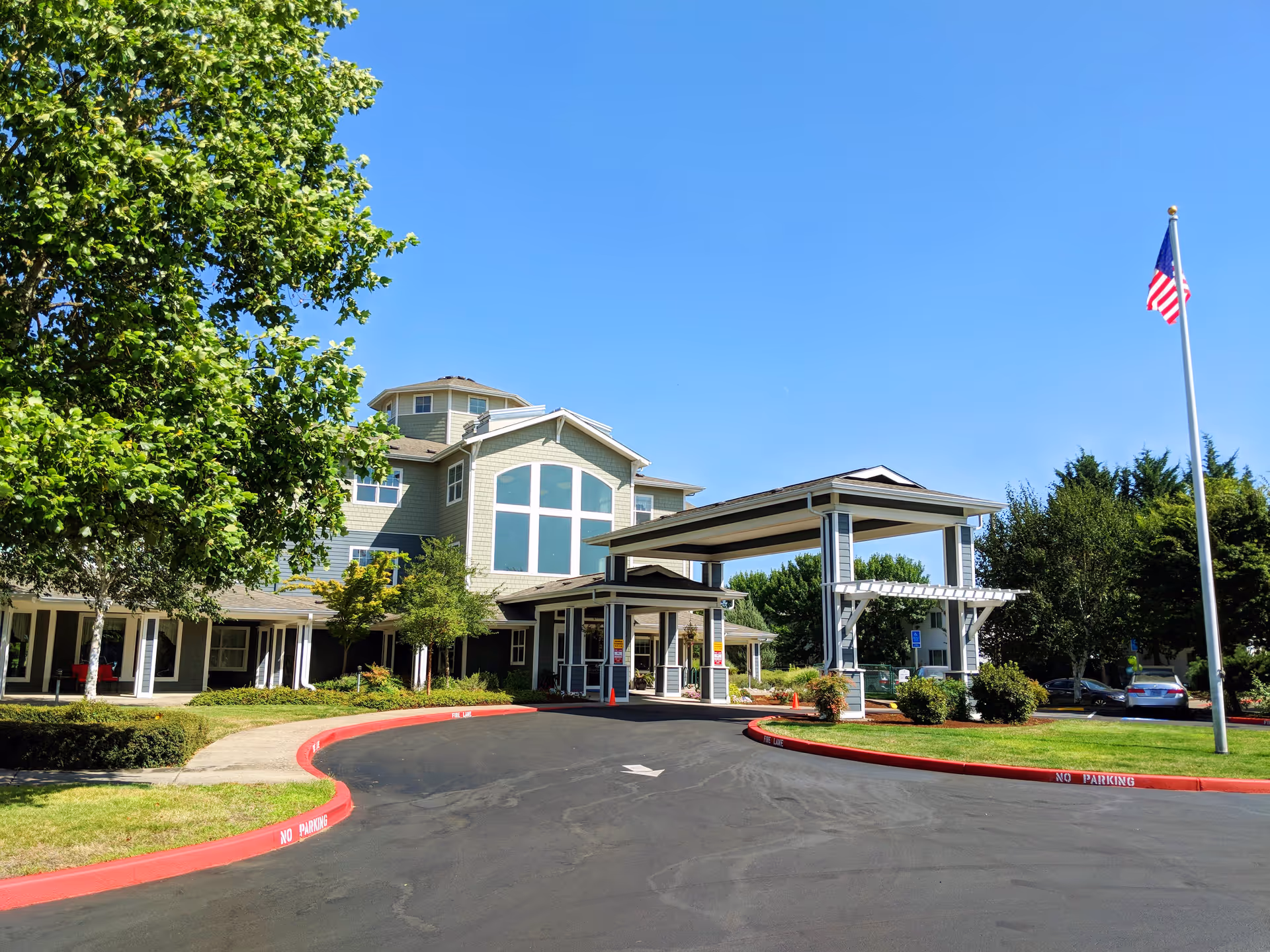 Exterior view of Timber Pointe Senior Living facility on a clear sunny day, showing the main entrance with a covered drop-off area, surrounded by green trees and a well-maintained lawn, with an American flag on a flagpole to the right.
