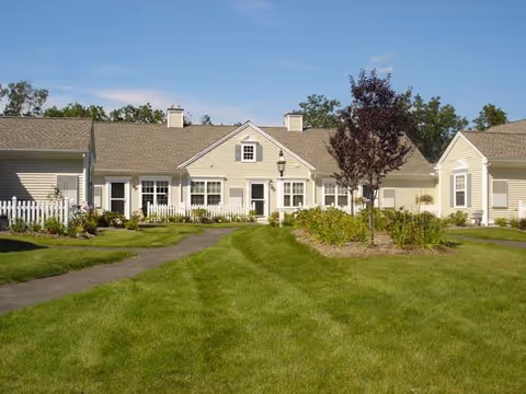 Single-story beige residential building with white trim and multiple windows, surrounded by a well-maintained green lawn, paved walkways, small trees, and shrubs under a clear blue sky.