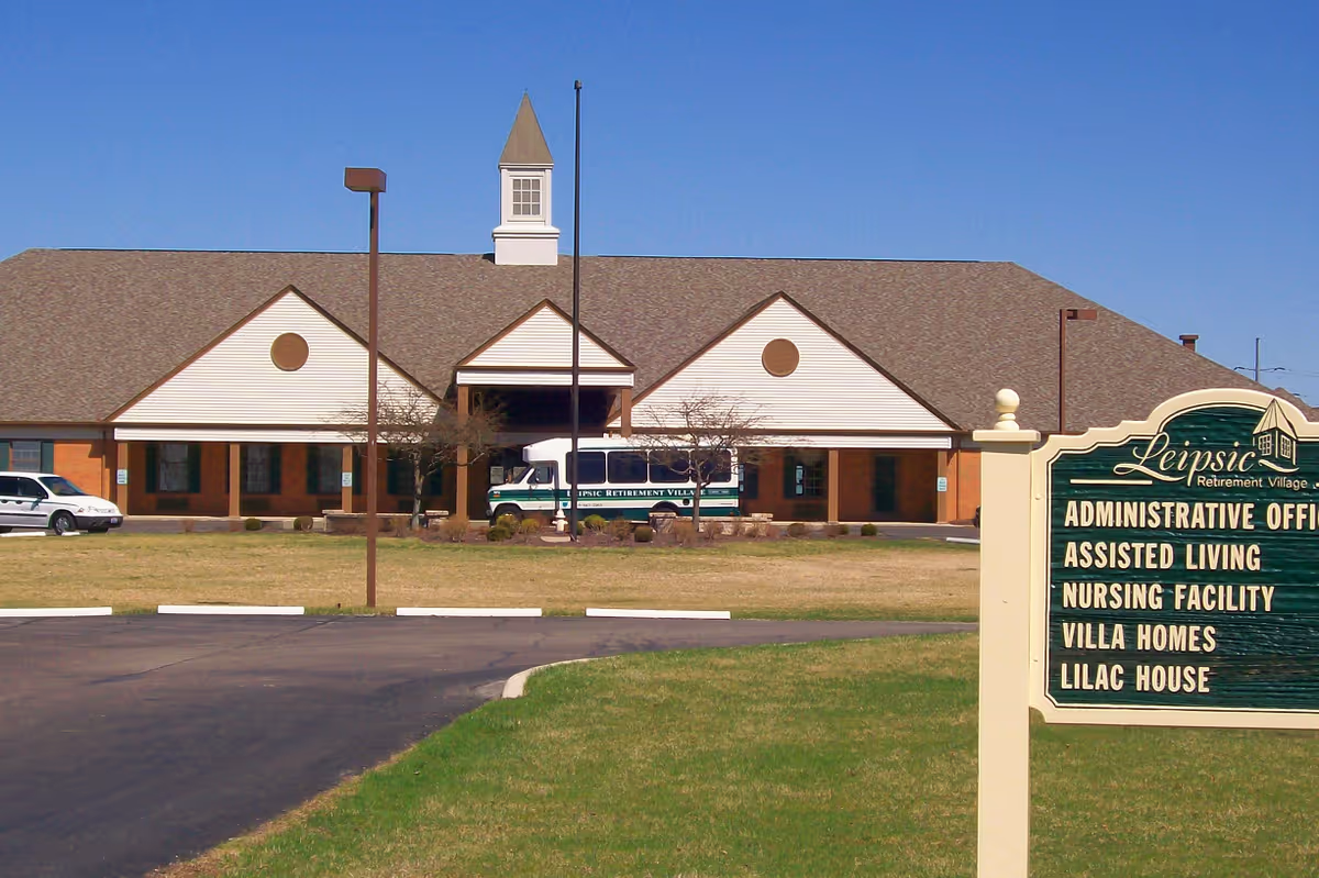 Front exterior of a single-story senior living facility with a lawn, parking area, shuttle van, and a large sign listing services.