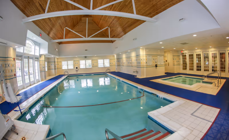 Indoor swimming pool and adjacent hot tub in a bright recreation room with a wooden vaulted ceiling and large windows.