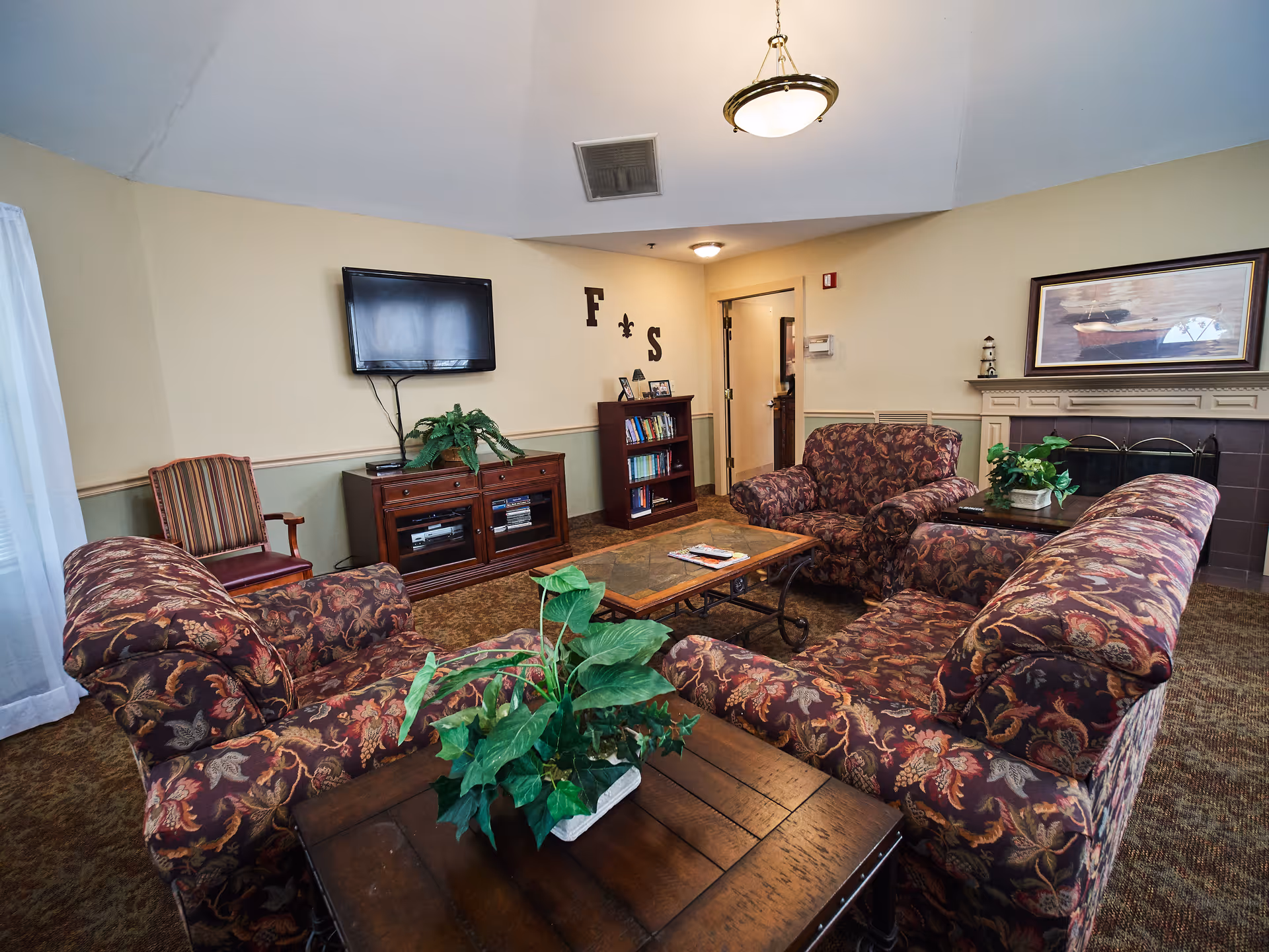 A cozy senior living facility common area with patterned upholstered sofas and armchairs arranged around a wooden coffee table. There is a TV mounted on the wall above a wooden cabinet, a small bookshelf, and a fireplace with a framed picture above it. The room is decorated with green plants and has warm lighting.