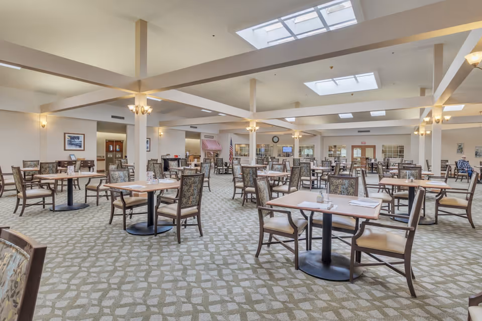 Spacious senior living dining room with multiple tables and chairs under skylights and soft lighting.
